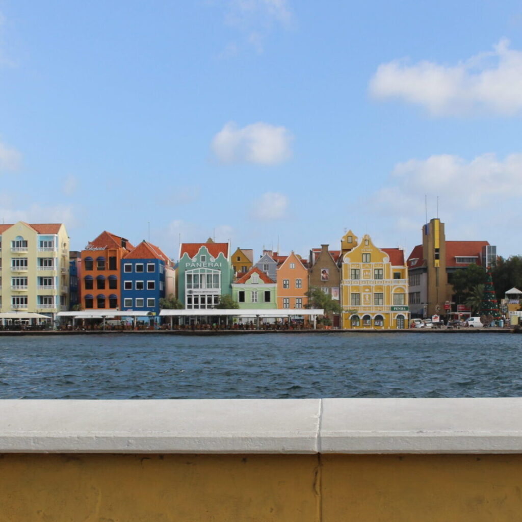 Image of the picturesque Handelskade in Willemstad, Curacao, featuring colorful historic buildings lining the waterfront under a clear blue sky.