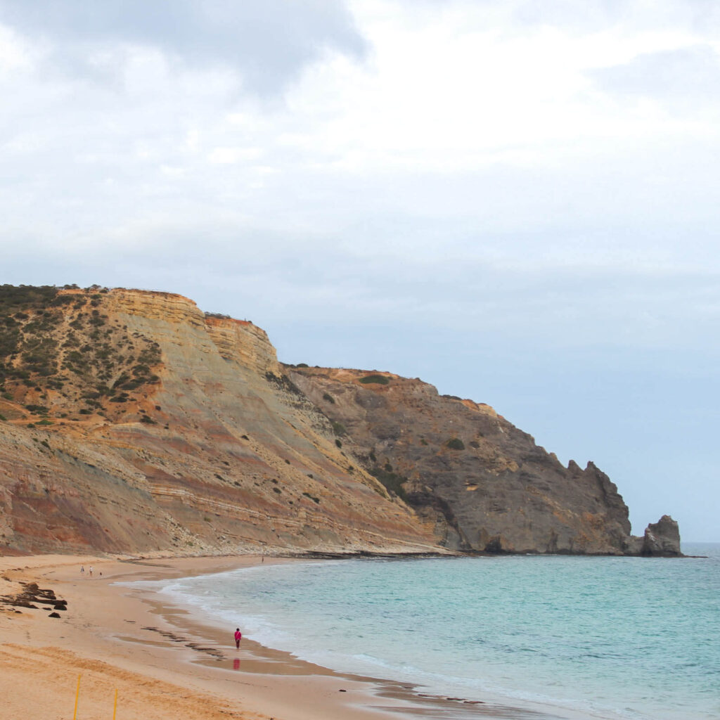 “Colorful layered cliffs rise beside the beach at Praia da Luz, with the ocean stretching out under a clear blue sky.”