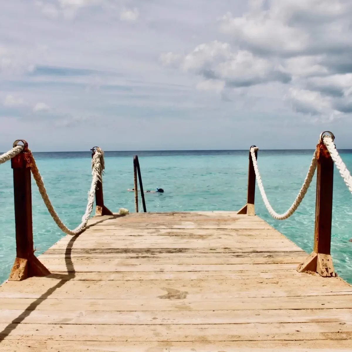 A wooden deck with rope handrails stretches out toward the azure blue sea at Kokomo Beach in Curaçao, surrounded by clear skies and tranquil waters.